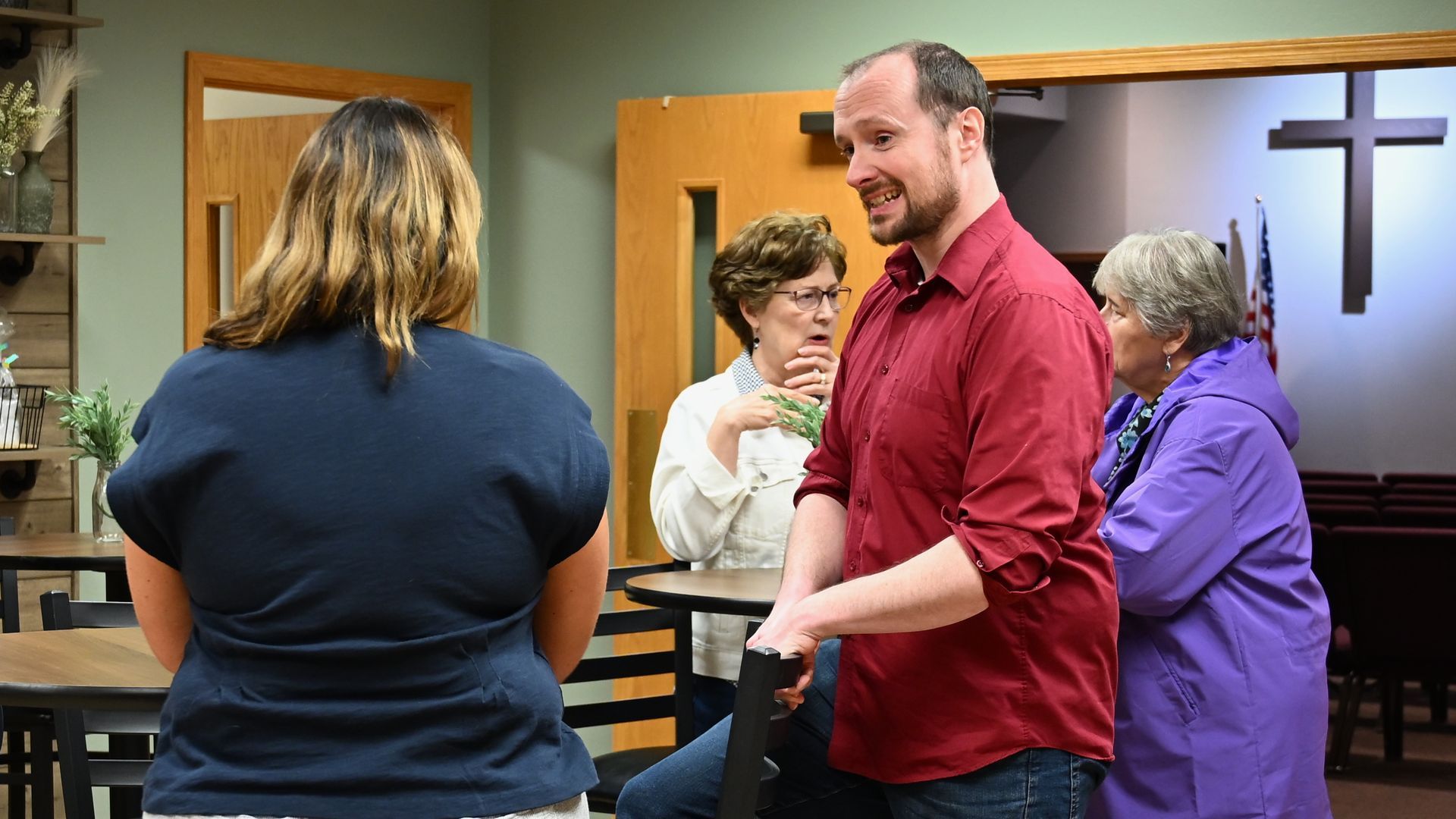 People talking in a church hall, with a red-shirted man speaking near a cross.