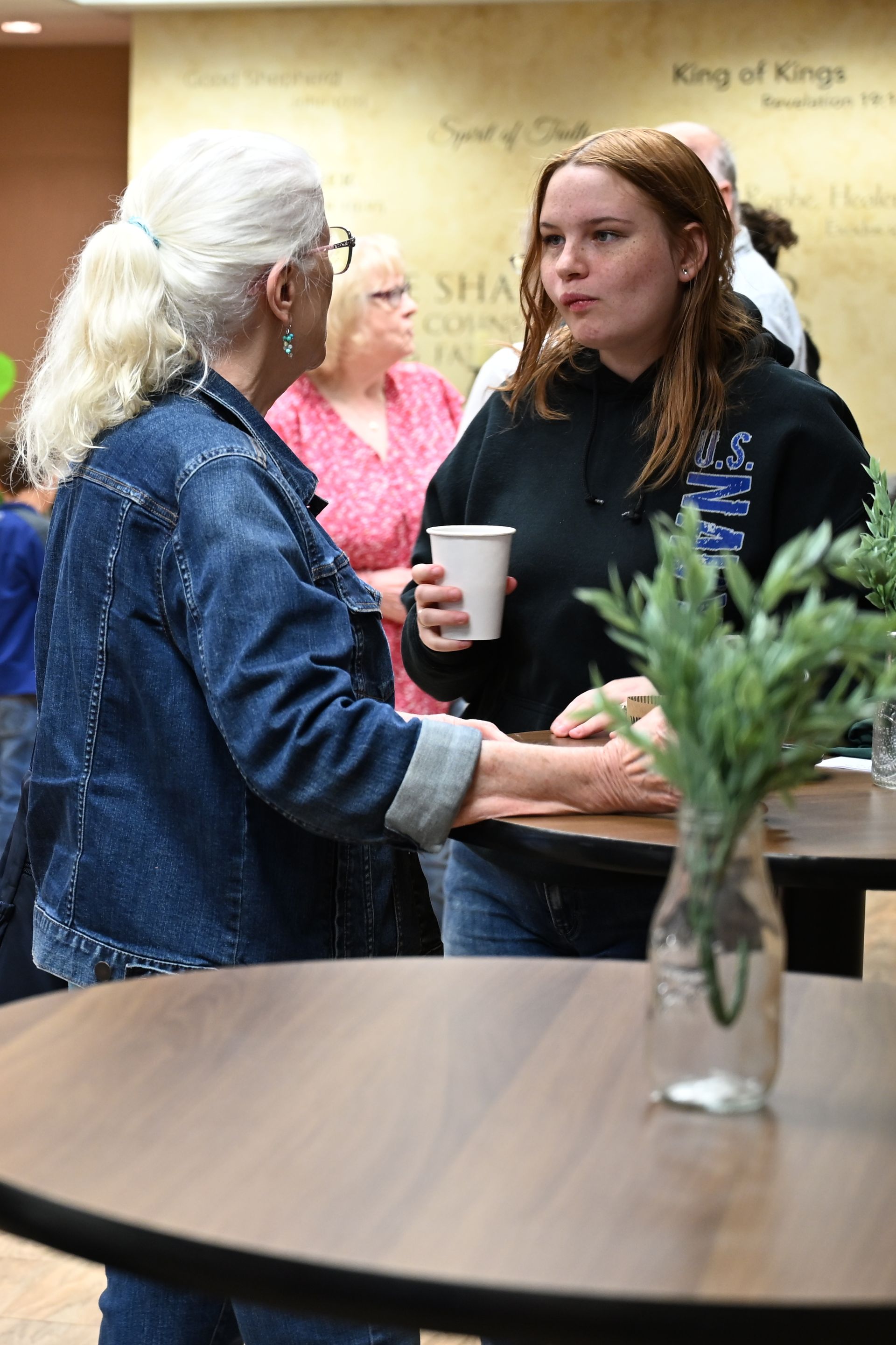 Two people talking at a café table, one holding a white cup, with a vase of greenery in the foreground.