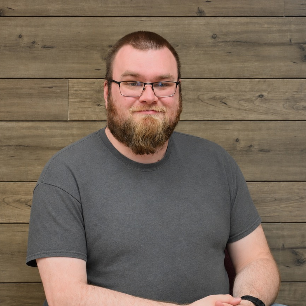 Man with glasses and beard in a gray T-shirt standing before a wooden plank wall