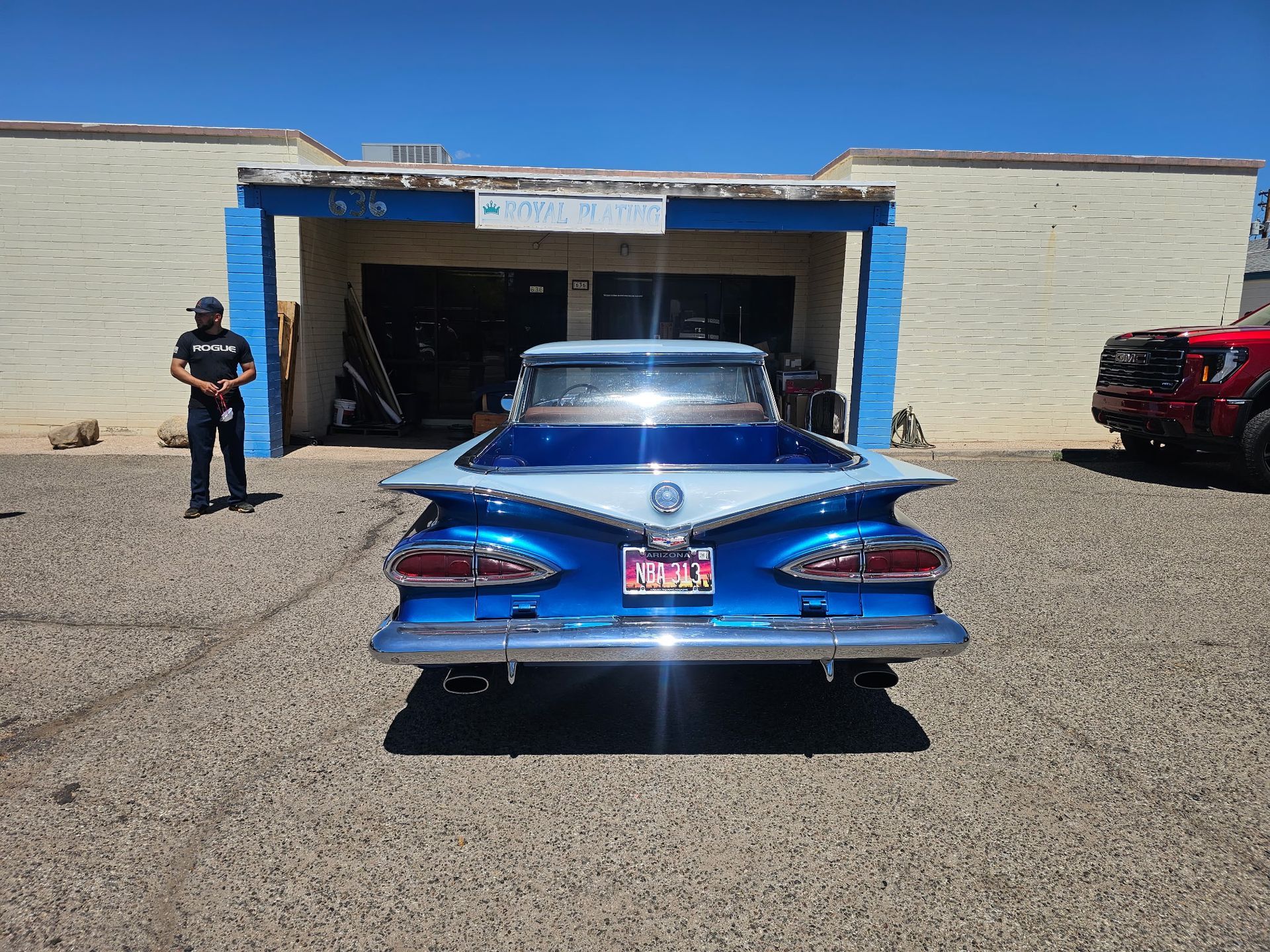 Blue classic car parked outside a garage, with a person standing to the left.