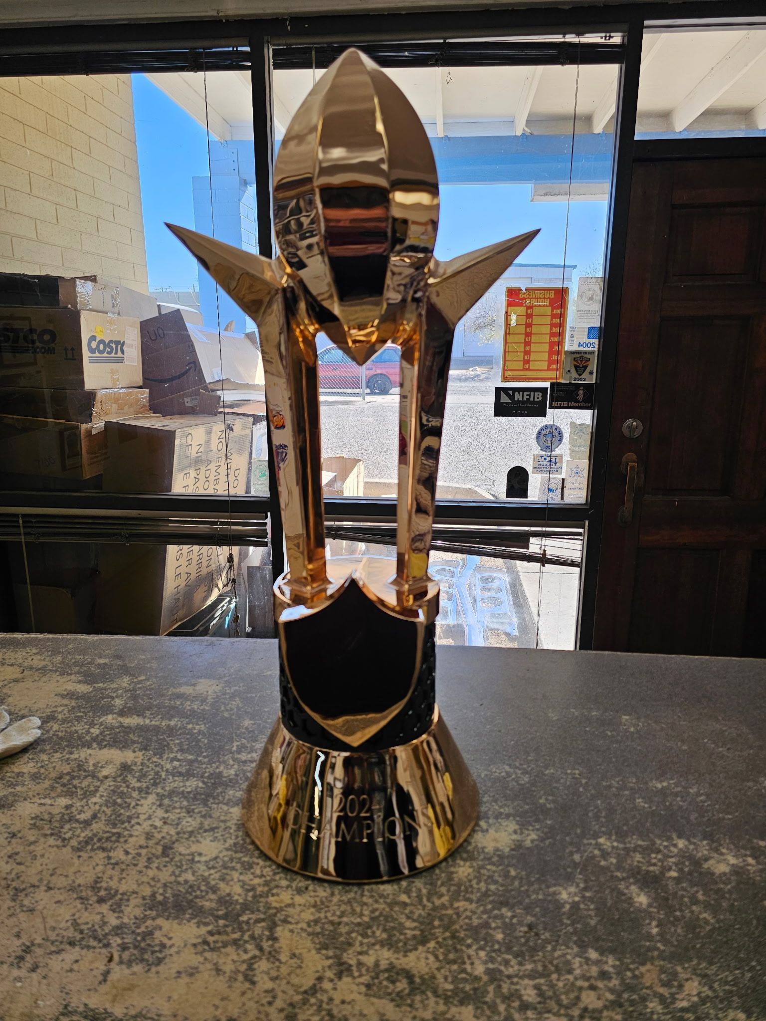 Gold trophy sculpture on a table, framed by a shop window with shelves and street view behind it
