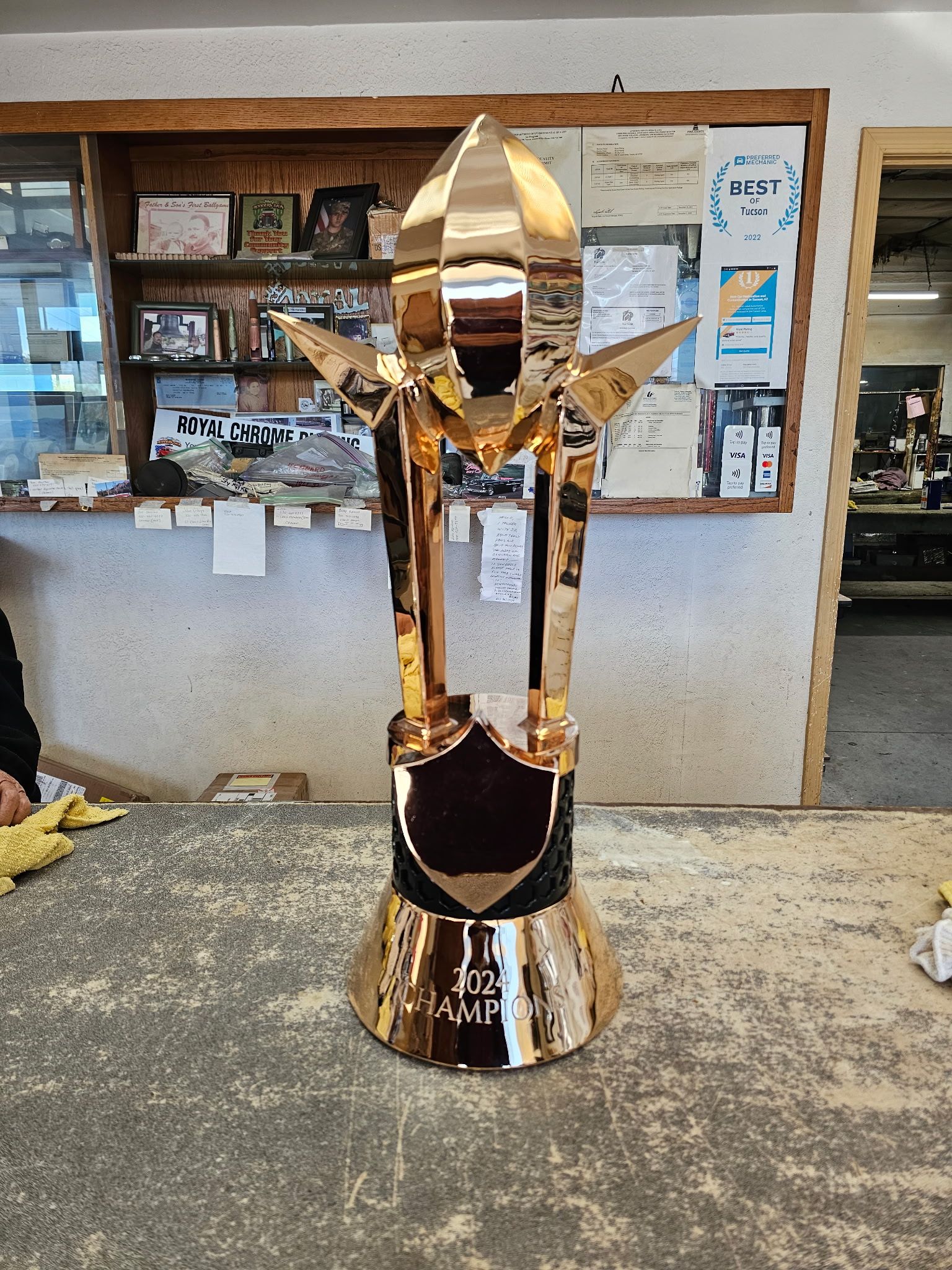Gold trophy statue on a worktable in a workshop, with shelves and posters in the background.