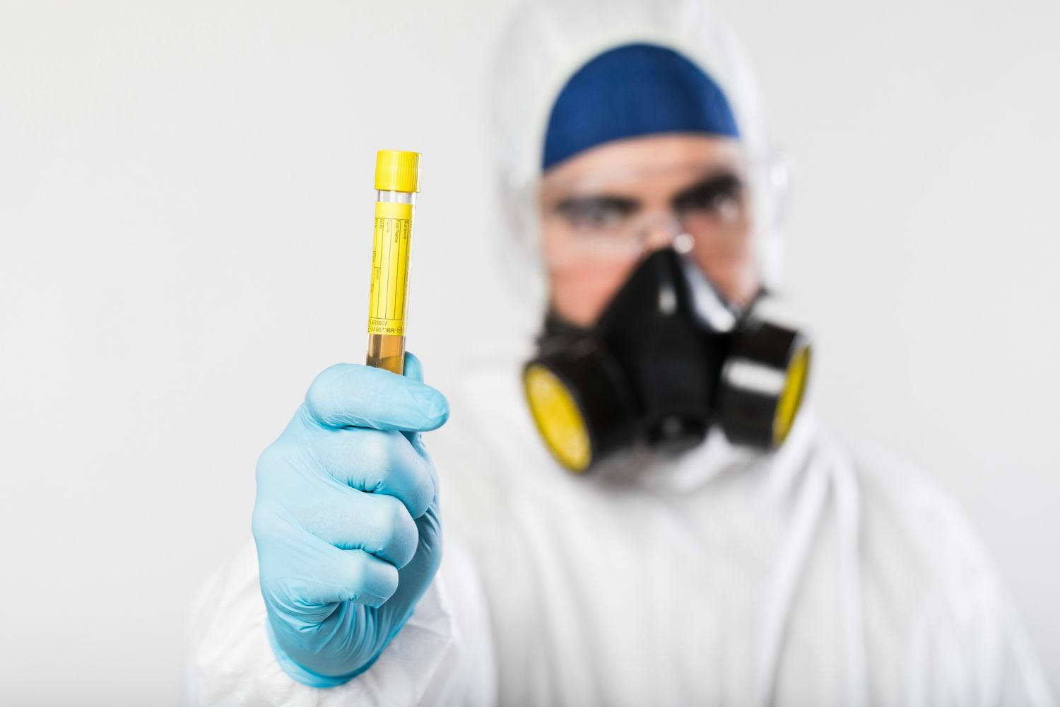 Gloved scientist holding a yellow test tube while wearing a respirator mask in a lab setting