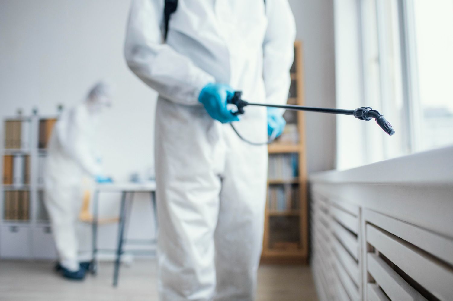 Worker in white protective suit spraying disinfectant along a wall indoors