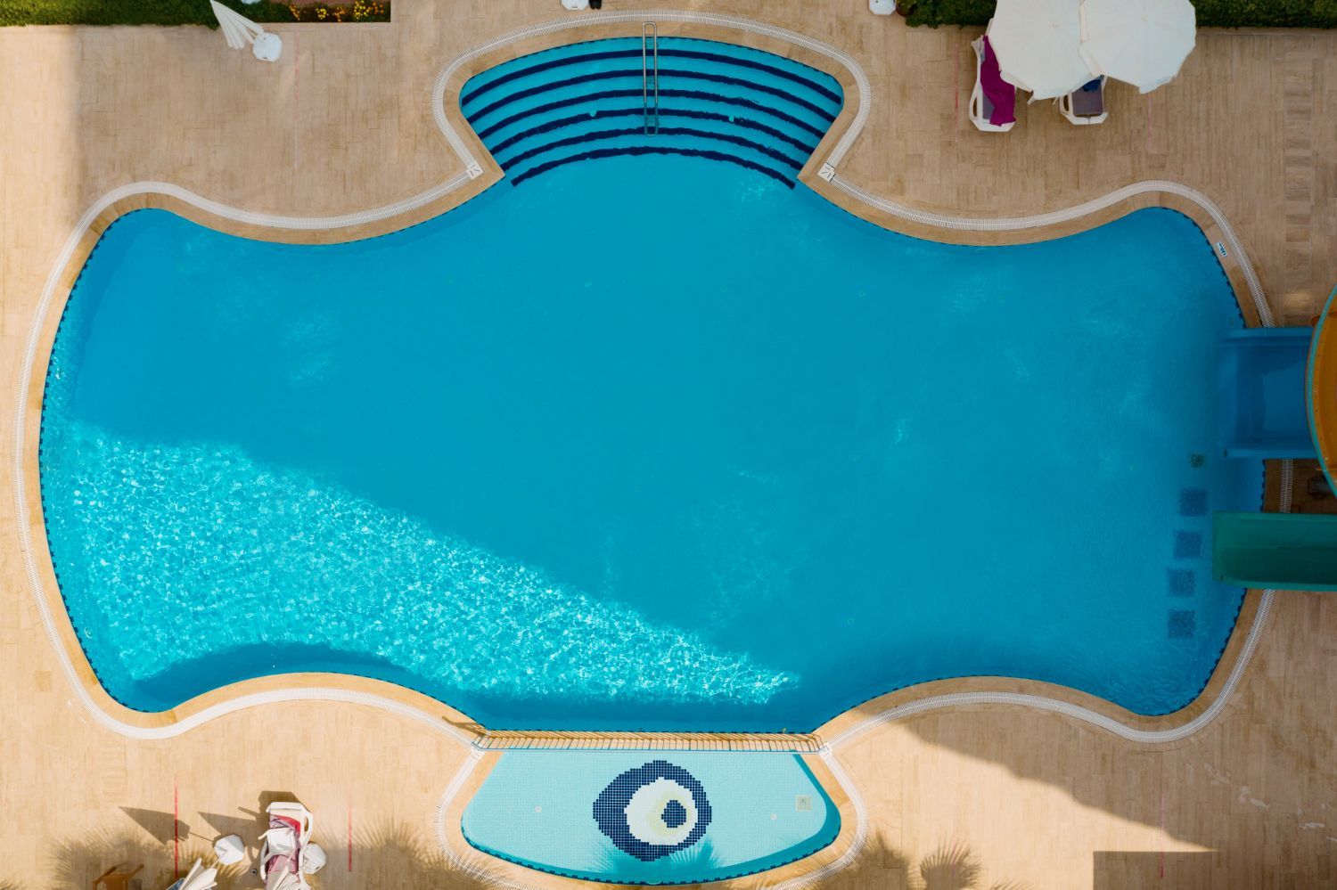 Overhead view of a blue swimming pool with curved edges, steps, and surrounding tan deck with umbrellas