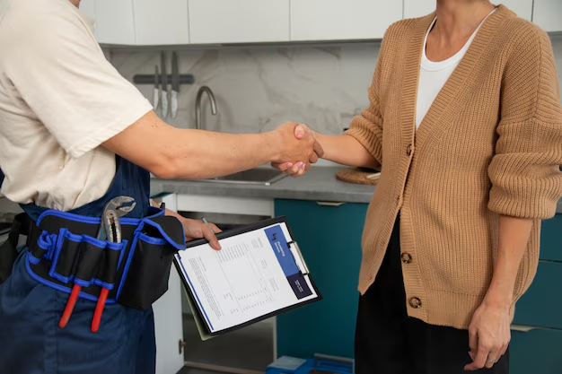 Two people shake hands in a kitchen, one wearing a tool belt and holding a clipboard.