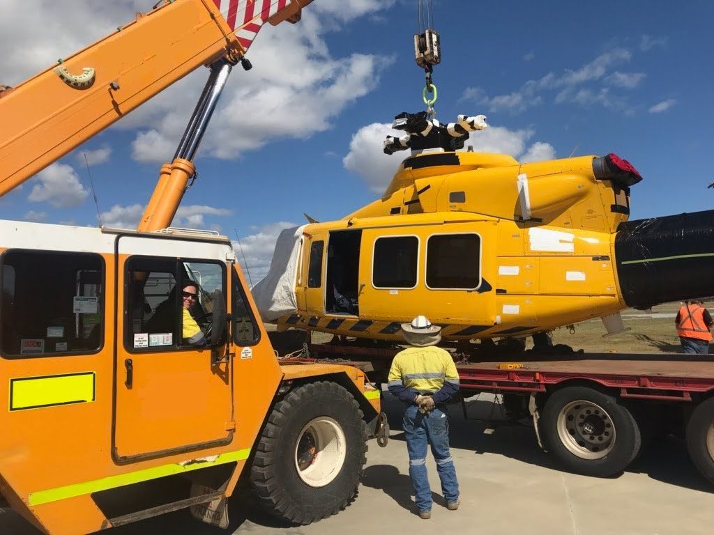 A crane lifts a yellow helicopter from a flatbed truck while a worker in high-visibility clothing stands nearby.
