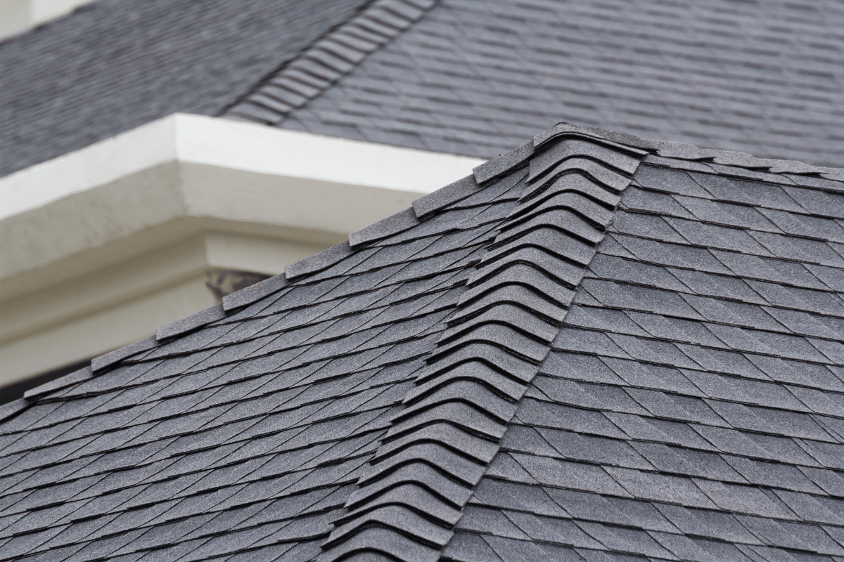 Gray shingled roof corner with white trim and a dormer window below a cloudy sky