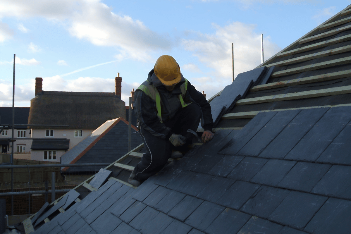 Worker in yellow hard hat repairing a slate roof on a sloped rooftop.