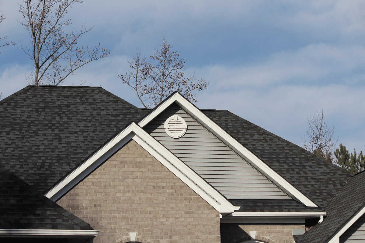 Suburban house roof with gray shingles, beige siding, and a circular attic vent under a cloudy sky