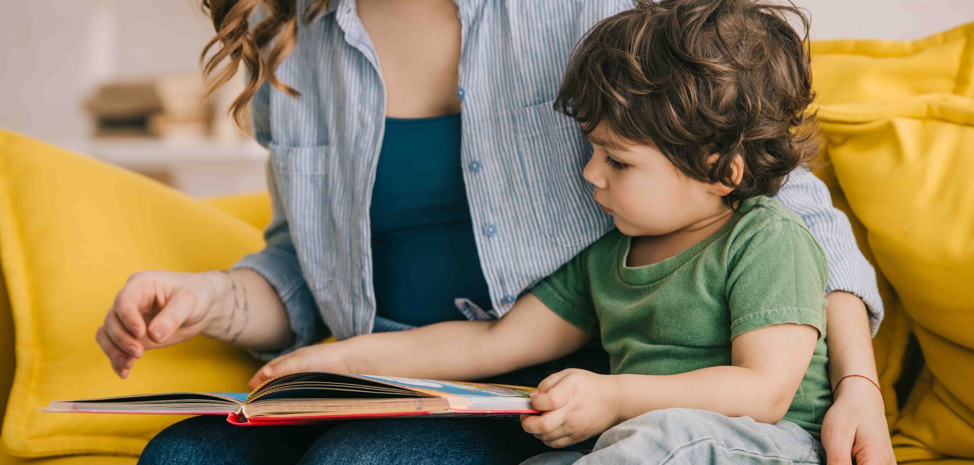 A woman is reading a book to a young boy while sitting on a yellow couch.
