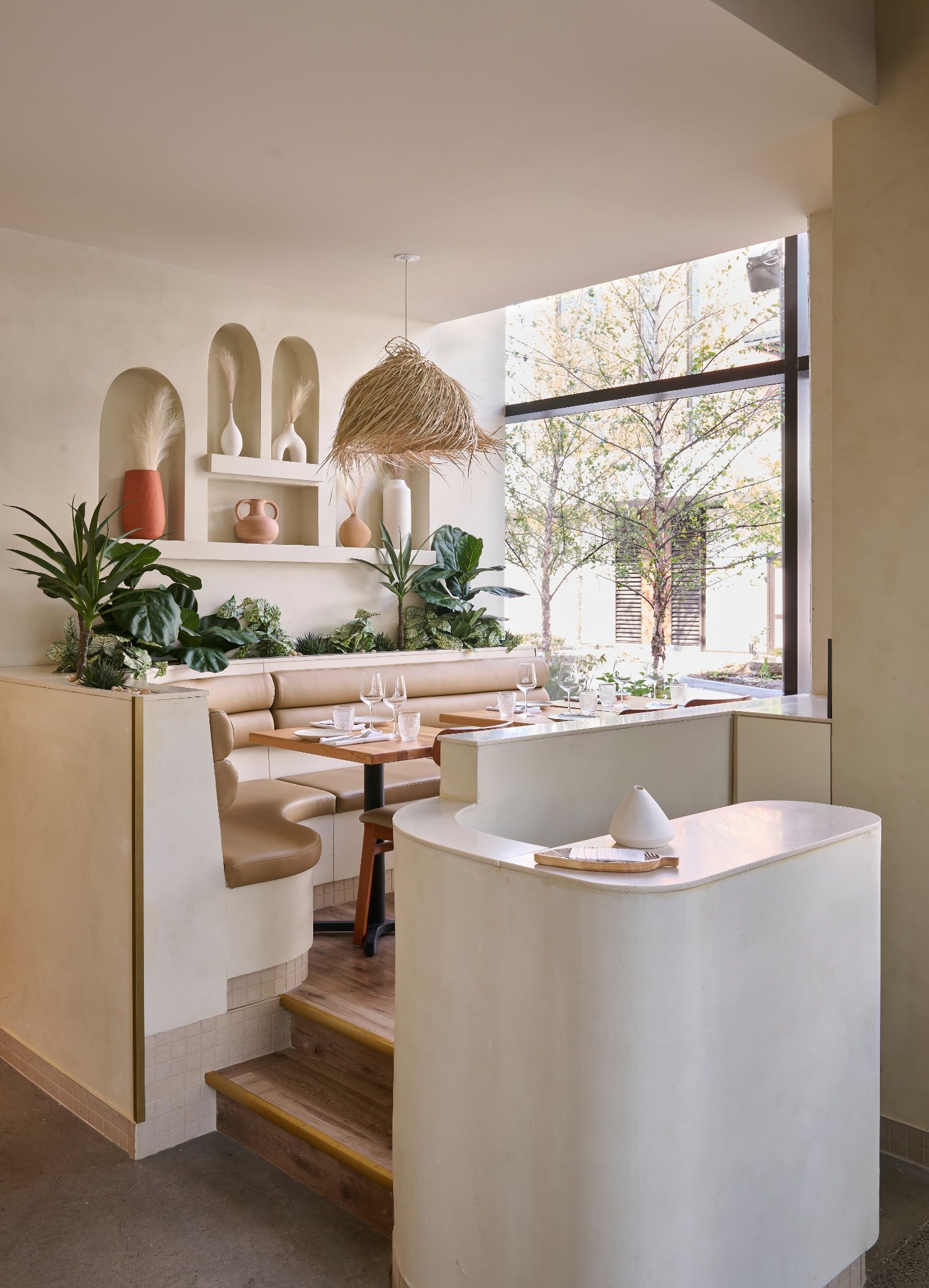 Intérieur lumineux de café avec tables en bois, plantes, étagères cintrées et une grande fenêtre près de l'espace salon.