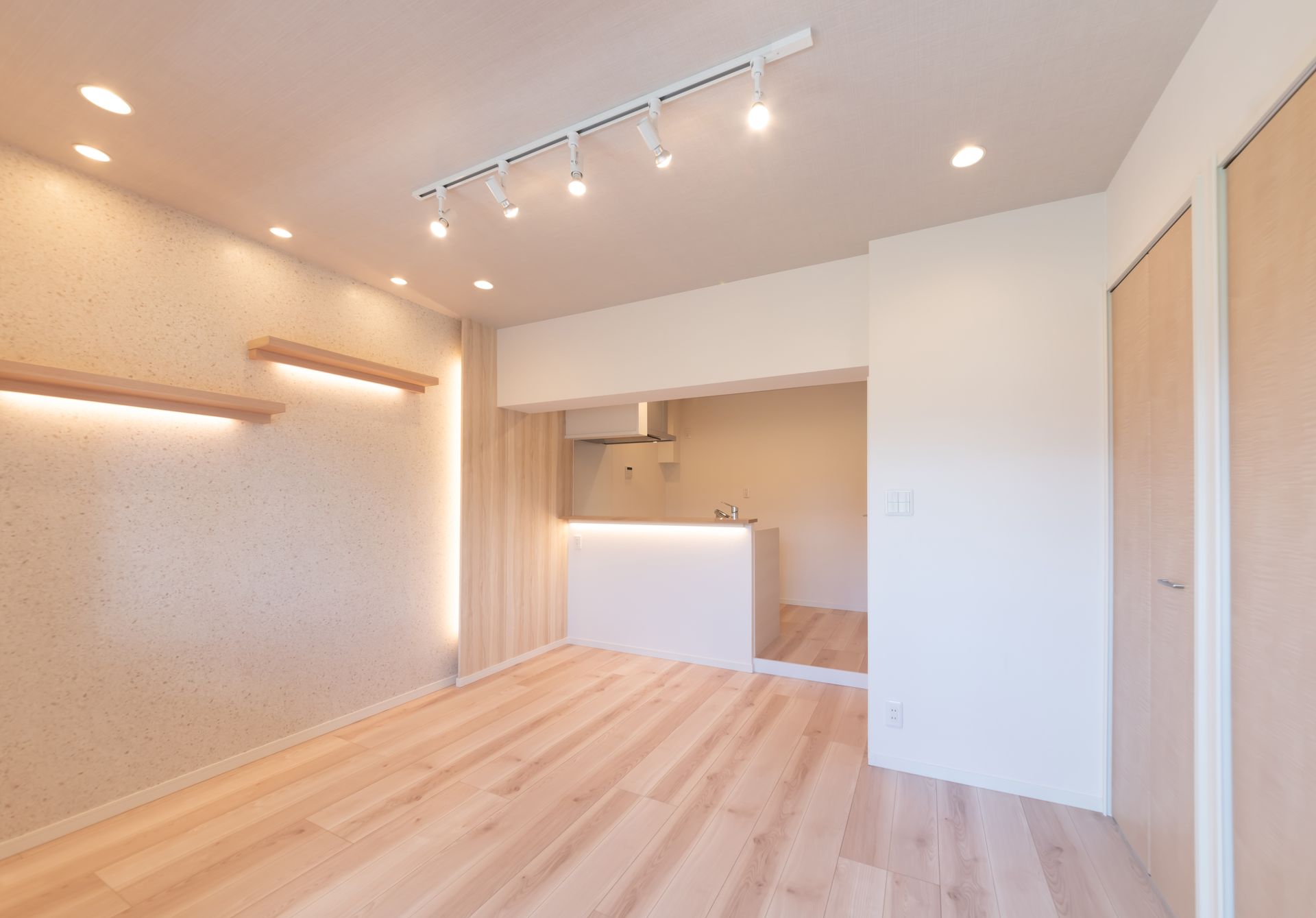 Pièce moderne vide avec parquet clair, murs blancs, spots encastrés au plafond et coin cuisine ouvert
