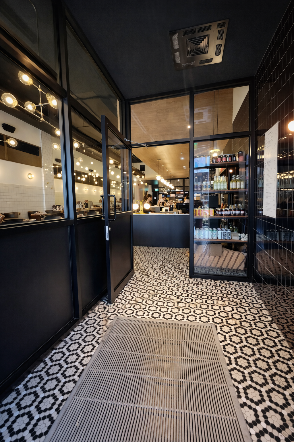 Entrée de café carrelée de noir avec portes vitrées, sol à motifs et éclairage intérieur chaleureux
