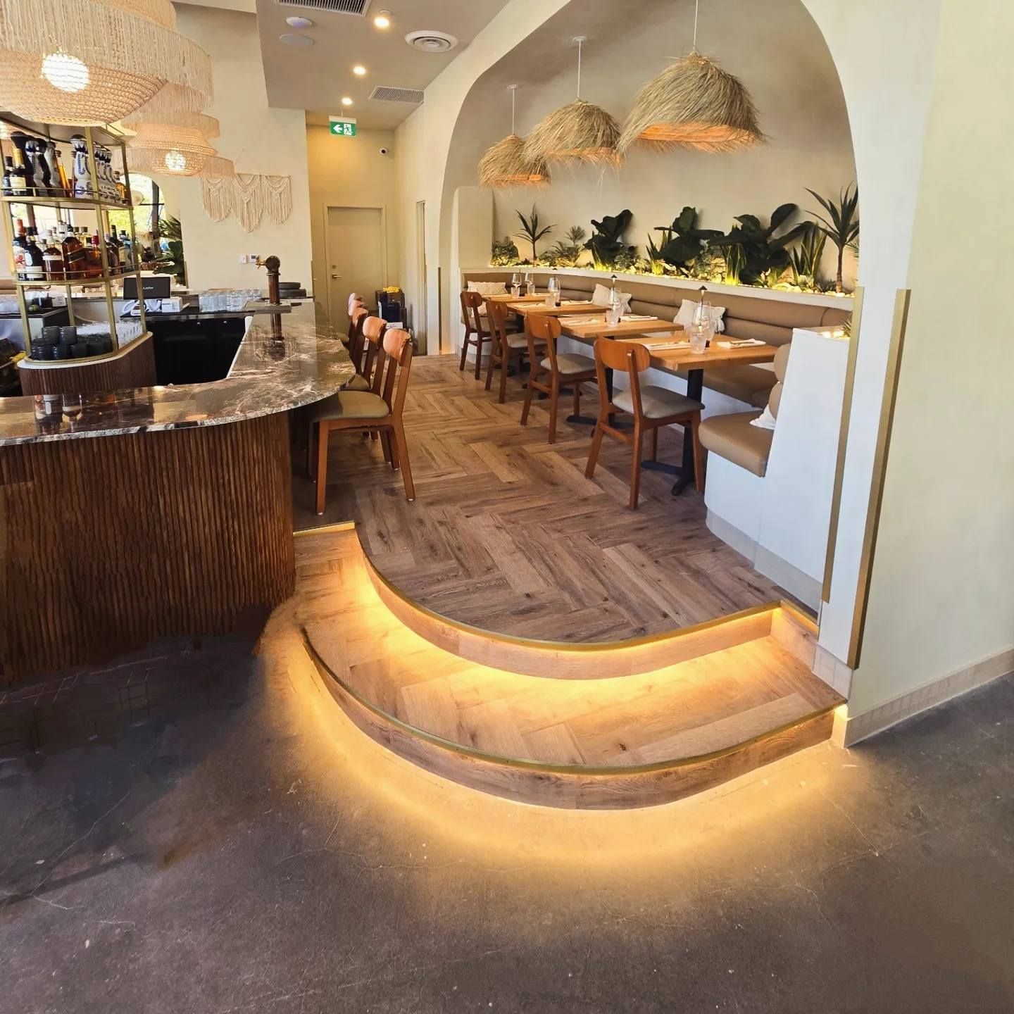 Intérieur de restaurant chaleureux avec une salle à manger surélevée et incurvée, des tables en bois, des suspensions lumineuses en tissu tressé et un éclairage tamisé.