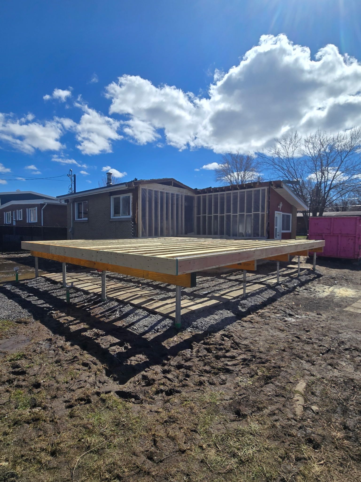 Plateforme en bois surélevée dans une cour boueuse sous un ciel d'un bleu éclatant, avec des maisons et une clôture en arrière-plan.