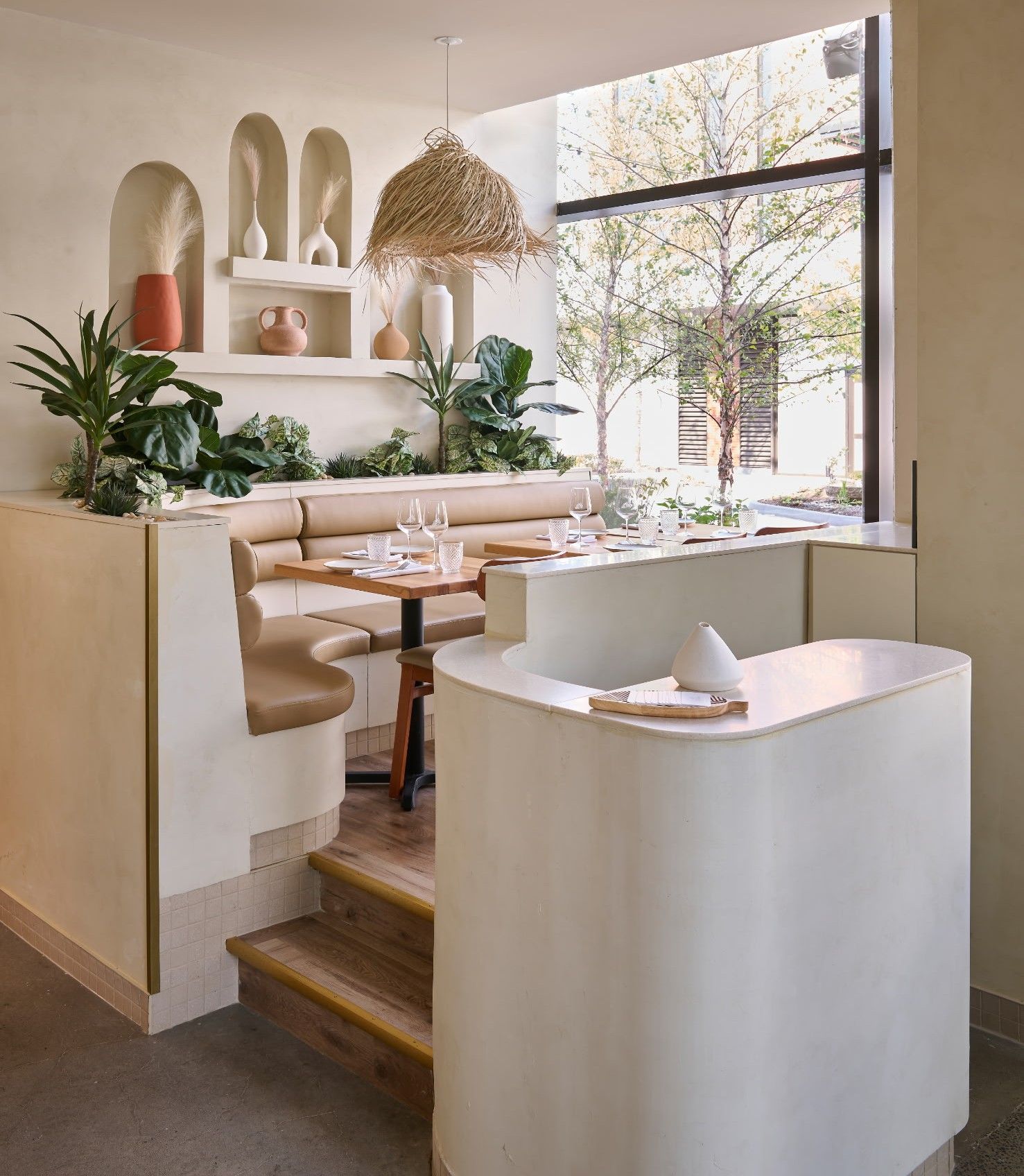 Café minimaliste et lumineux avec des sièges blancs incurvés, des touches de bois, des plantes et une grande fenêtre baignée de lumière naturelle.