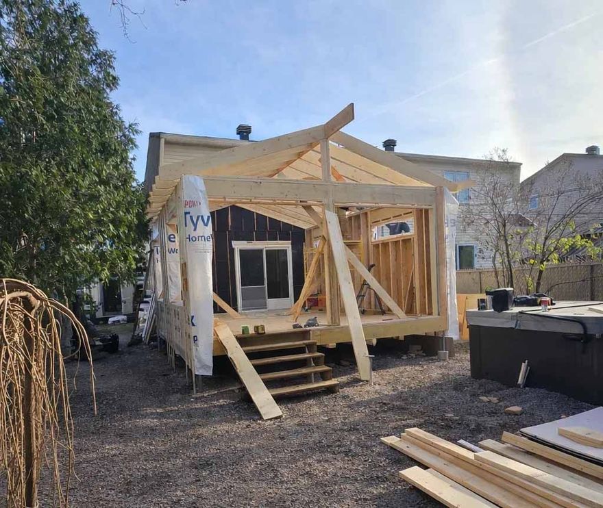 Charpente de porche en bois partiellement construite, adossée à une petite maison dans une cour, avec des marches et du bois de construction à proximité.