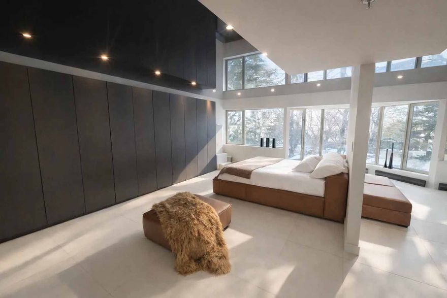 Chambre moderne avec baies vitrées, lambris foncés, linge de lit blanc et fauteuil beige sur un tapis clair.