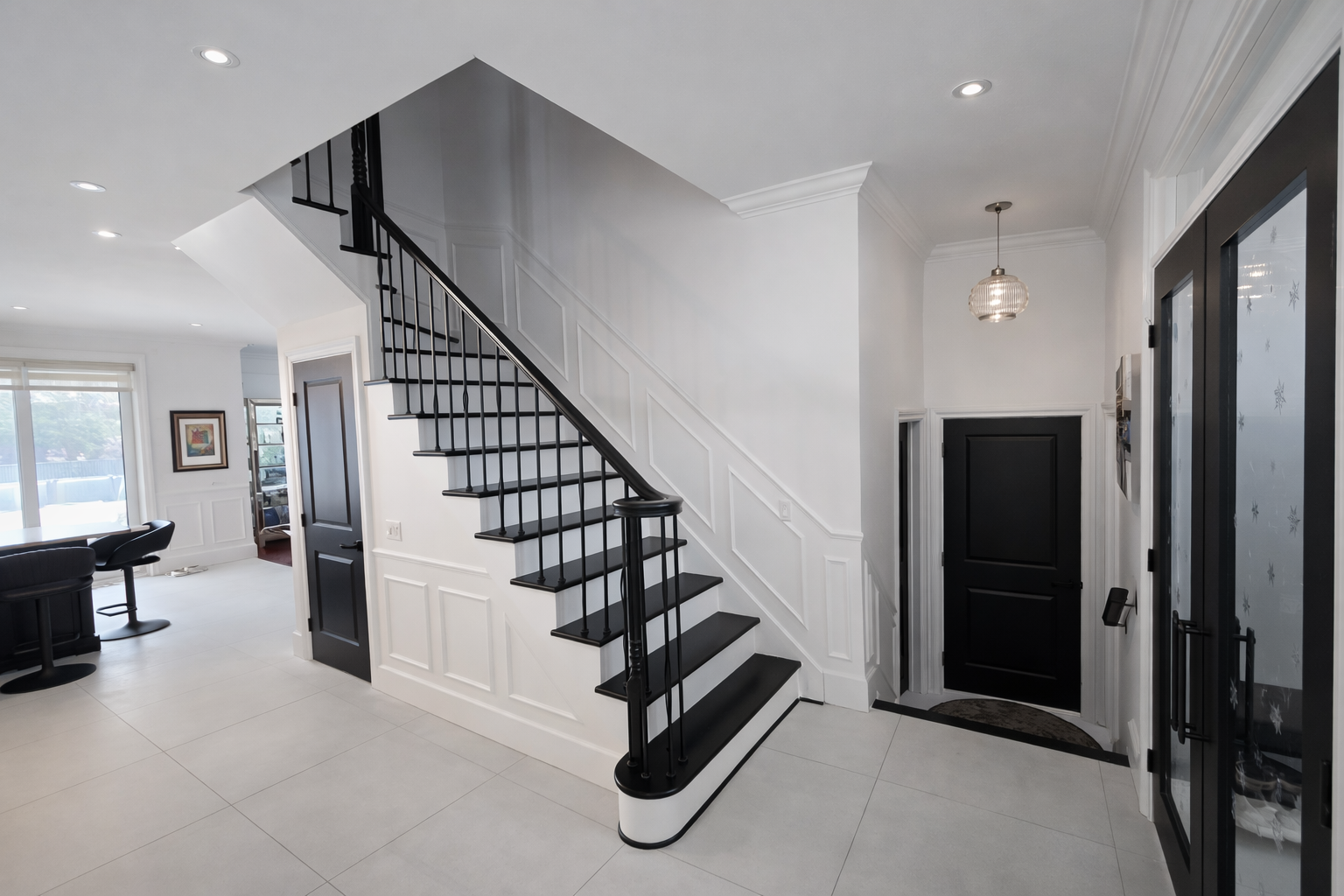 Hall d'entrée lumineux avec escalier noir, murs blancs et sol carrelé menant à une porte d'entrée
