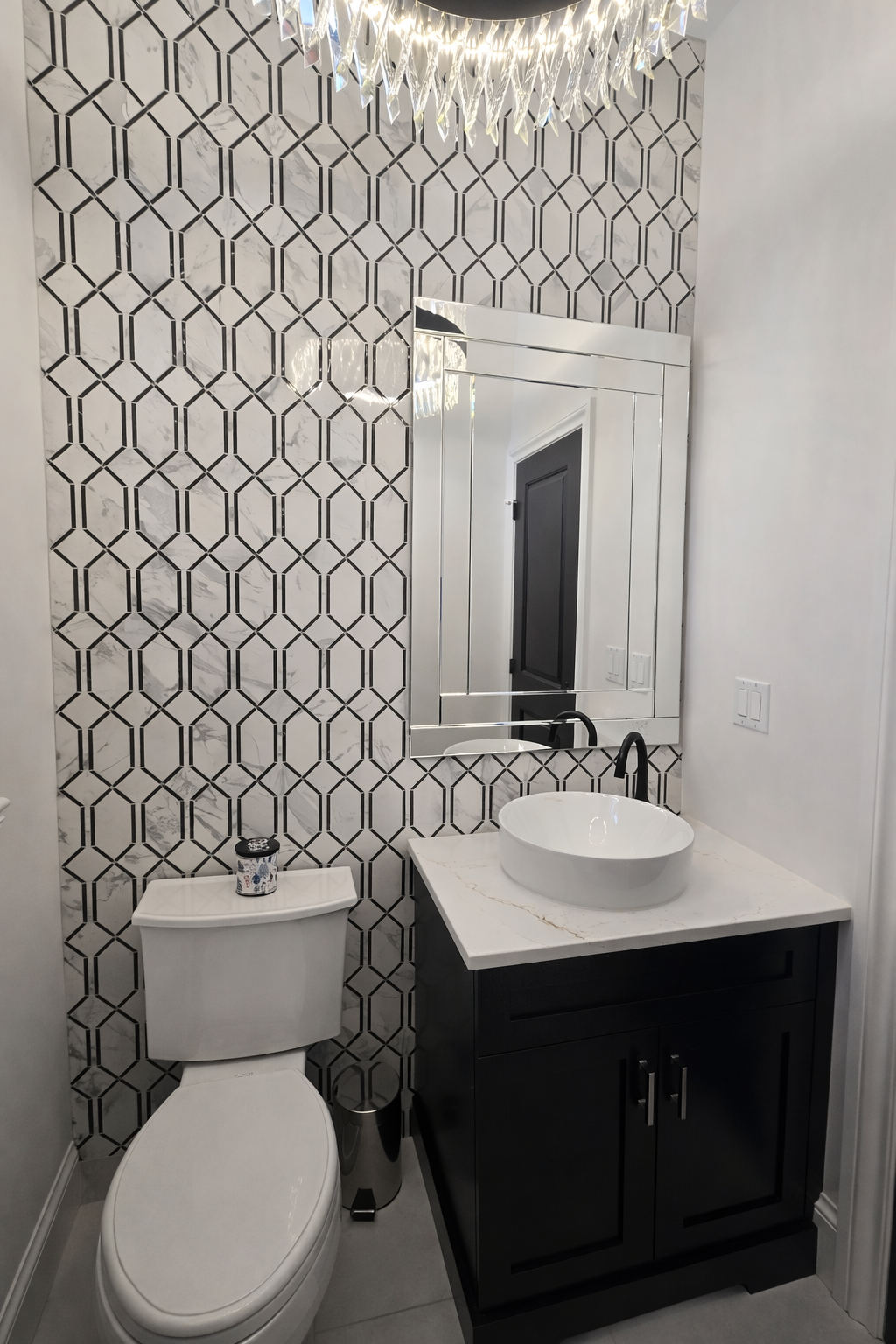 Petite salle de bains avec toilettes blanches, meuble-lavabo foncé, lavabo rond, armoire à miroir et mur d'accent en carrelage à motifs.