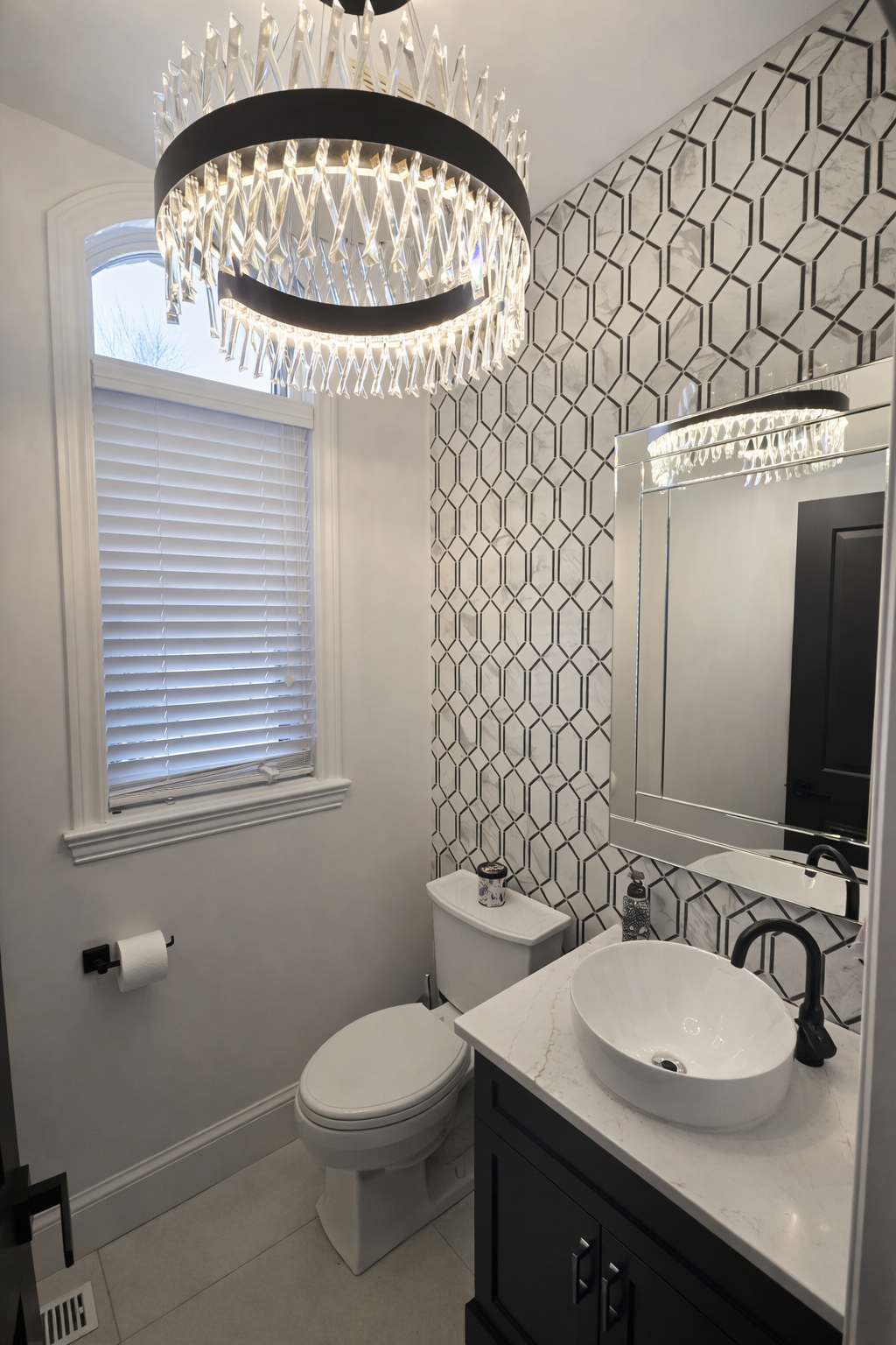 Petite salle de bains moderne avec papier peint à motifs, miroir rond, vasque, toilettes et lustre.