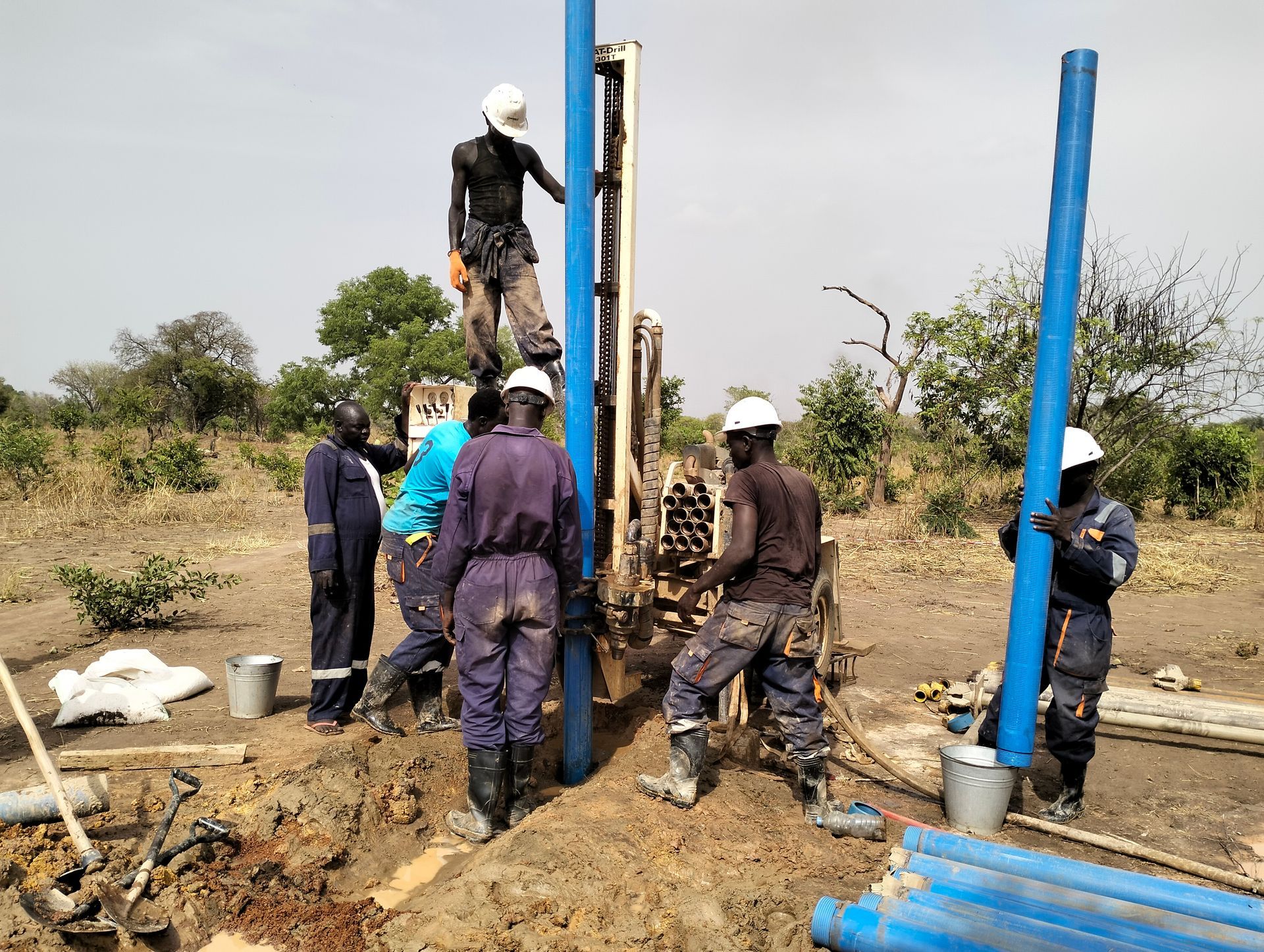 Workers drilling a borehole beside blue pipes in a muddy field