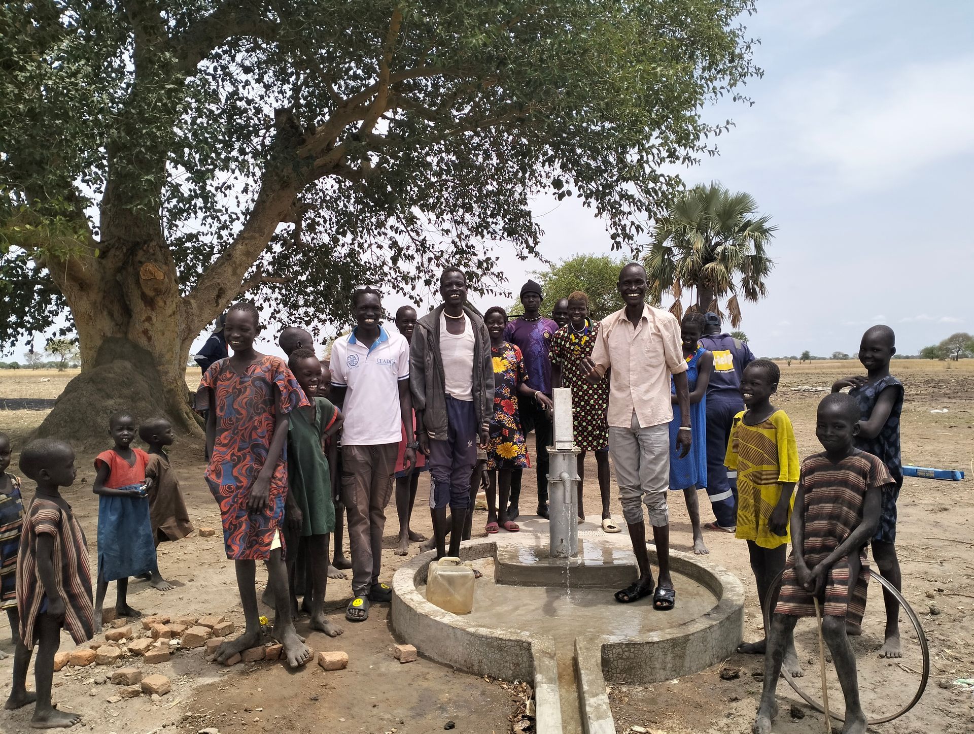 Group of people gathered around a hand pump and water point under a tree in a rural area