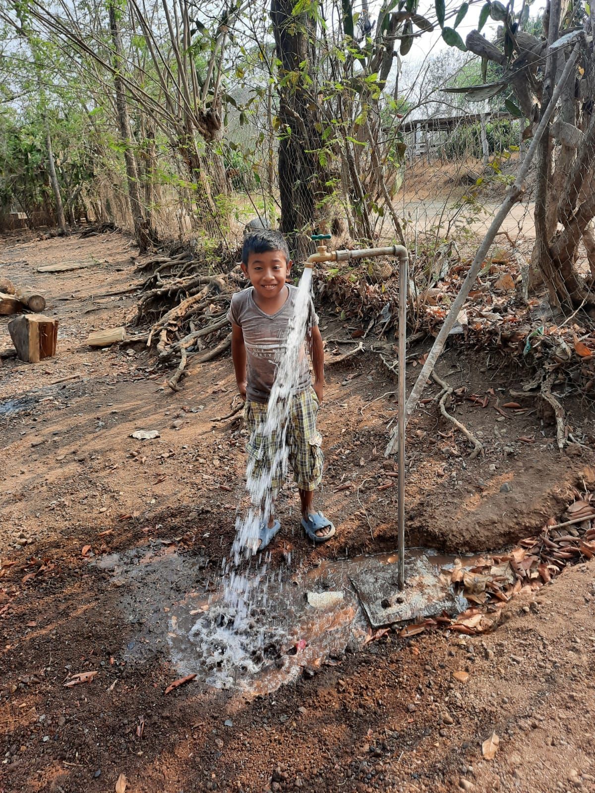 Boy standing in a dry wooded area, holding a stream of water from a hose or pipe onto the ground