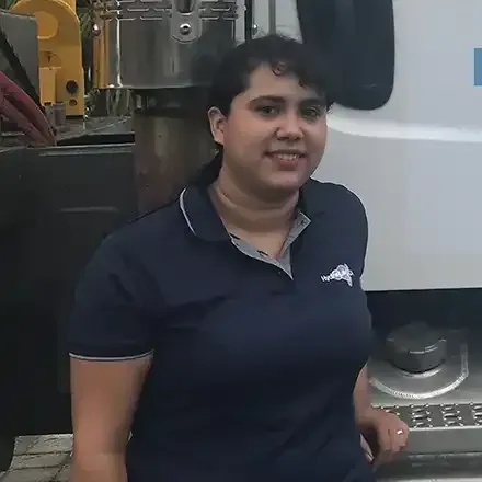 Person in a navy polo shirt standing beside industrial equipment and a machine panel, smiling at the camera.