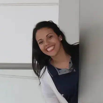 Smiling woman peeking from behind a white wall in a dark jacket and gray-collared shirt.