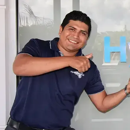 Smiling man in a navy polo points to a blue letter H on a glass wall.