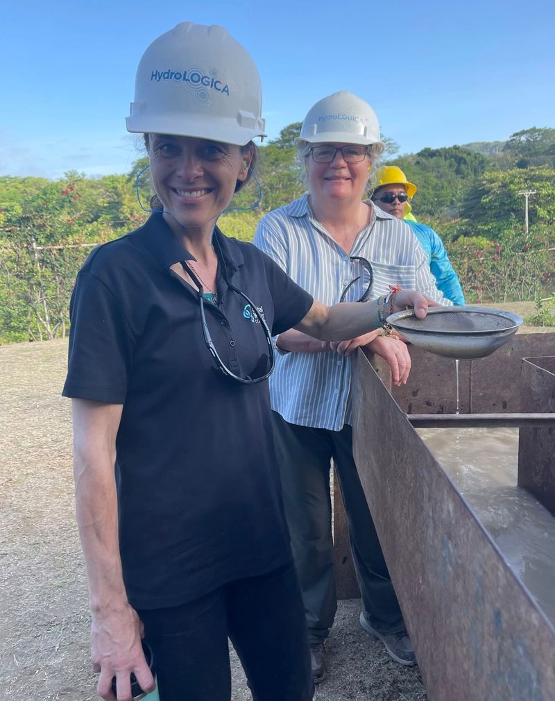 Three people in hard hats at a construction site, smiling by a concrete barrier with green hills behind them