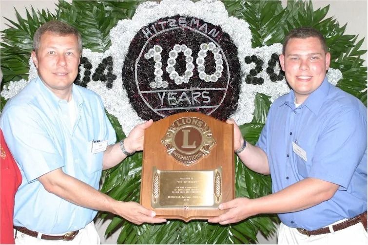 Two men holding an award plaque in front of a flower arrangement with 