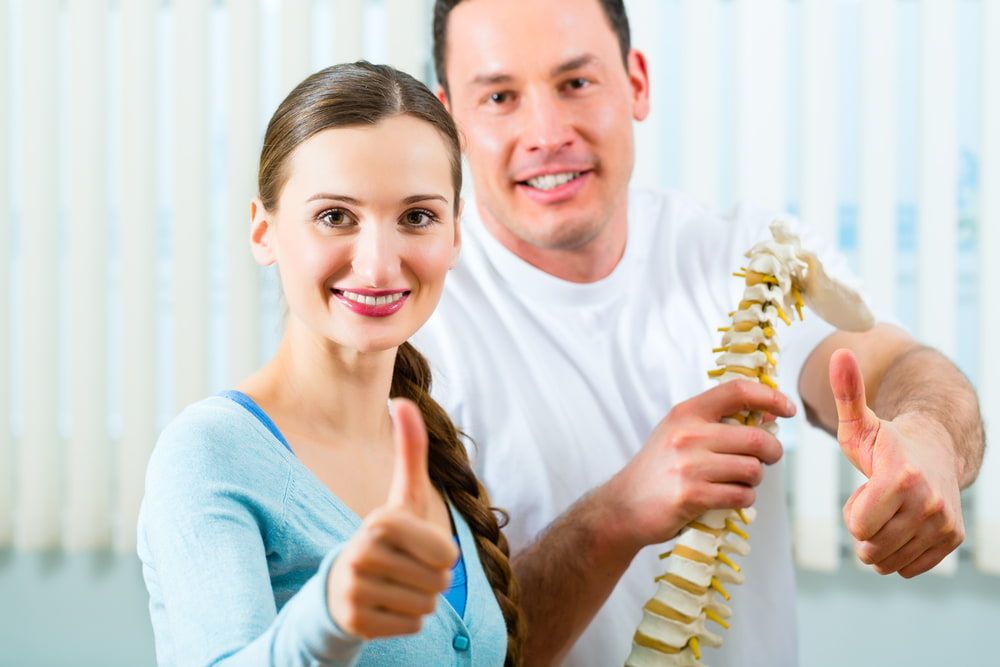 A Man And A Woman Are Giving A Thumbs Up While Holding A Model Of A Spine — Southern Districts Physiotherapy & Sports Injuries Clinic In Albion Park, NSW