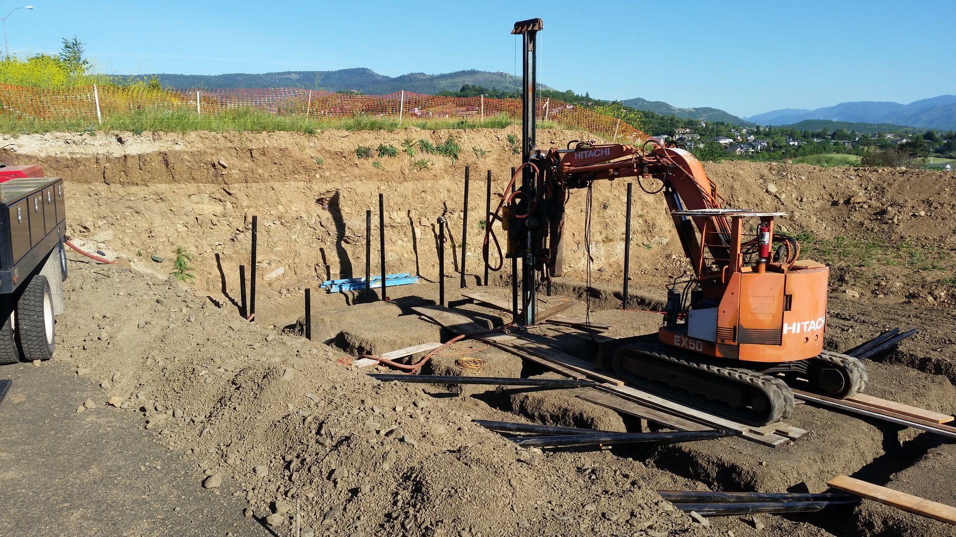 Construction site with excavator drilling into a hillside for retaining wall posts; sunny day.