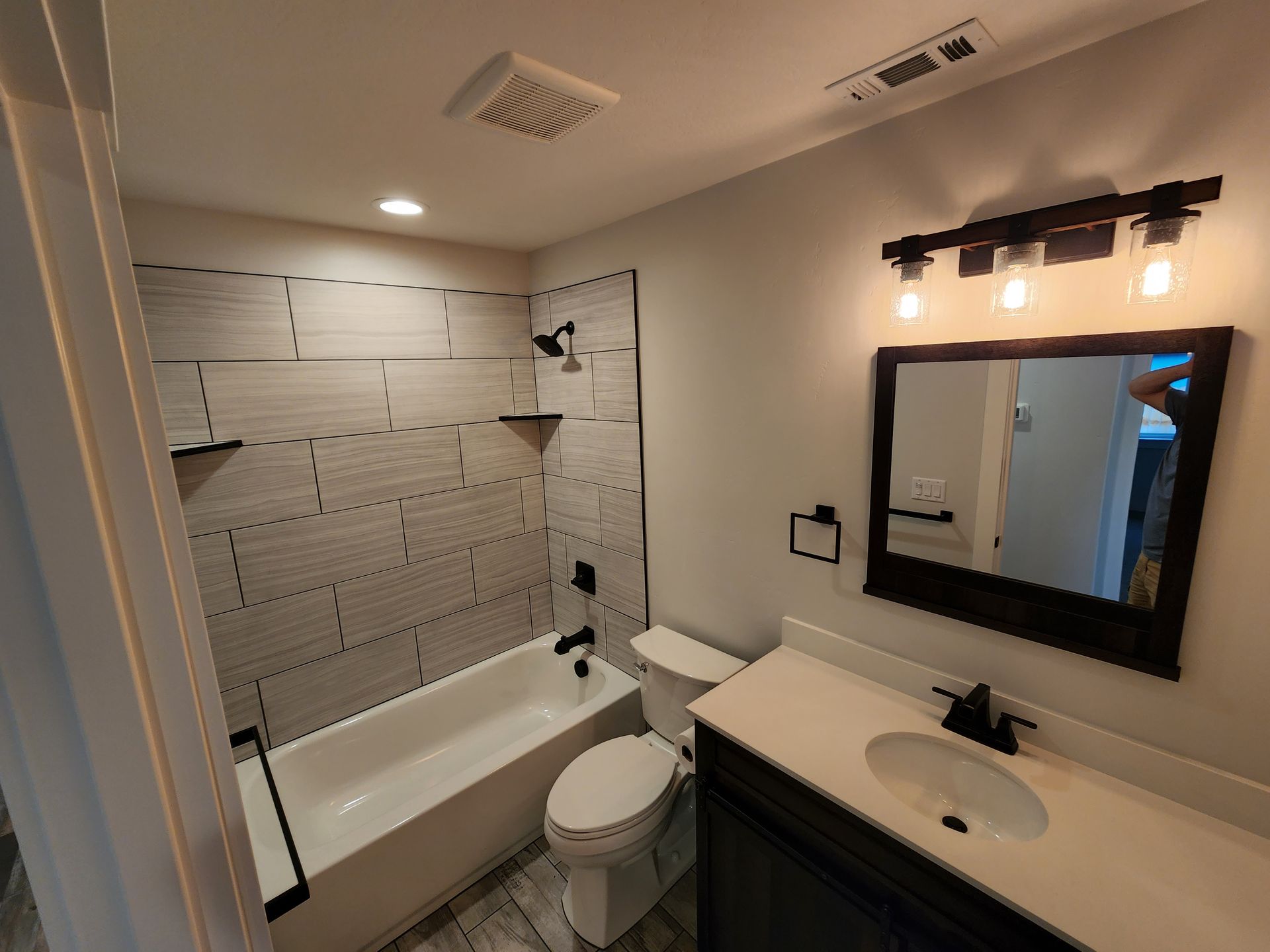 Bathroom with gray tile, white tub, vanity, black fixtures, and a mirror.