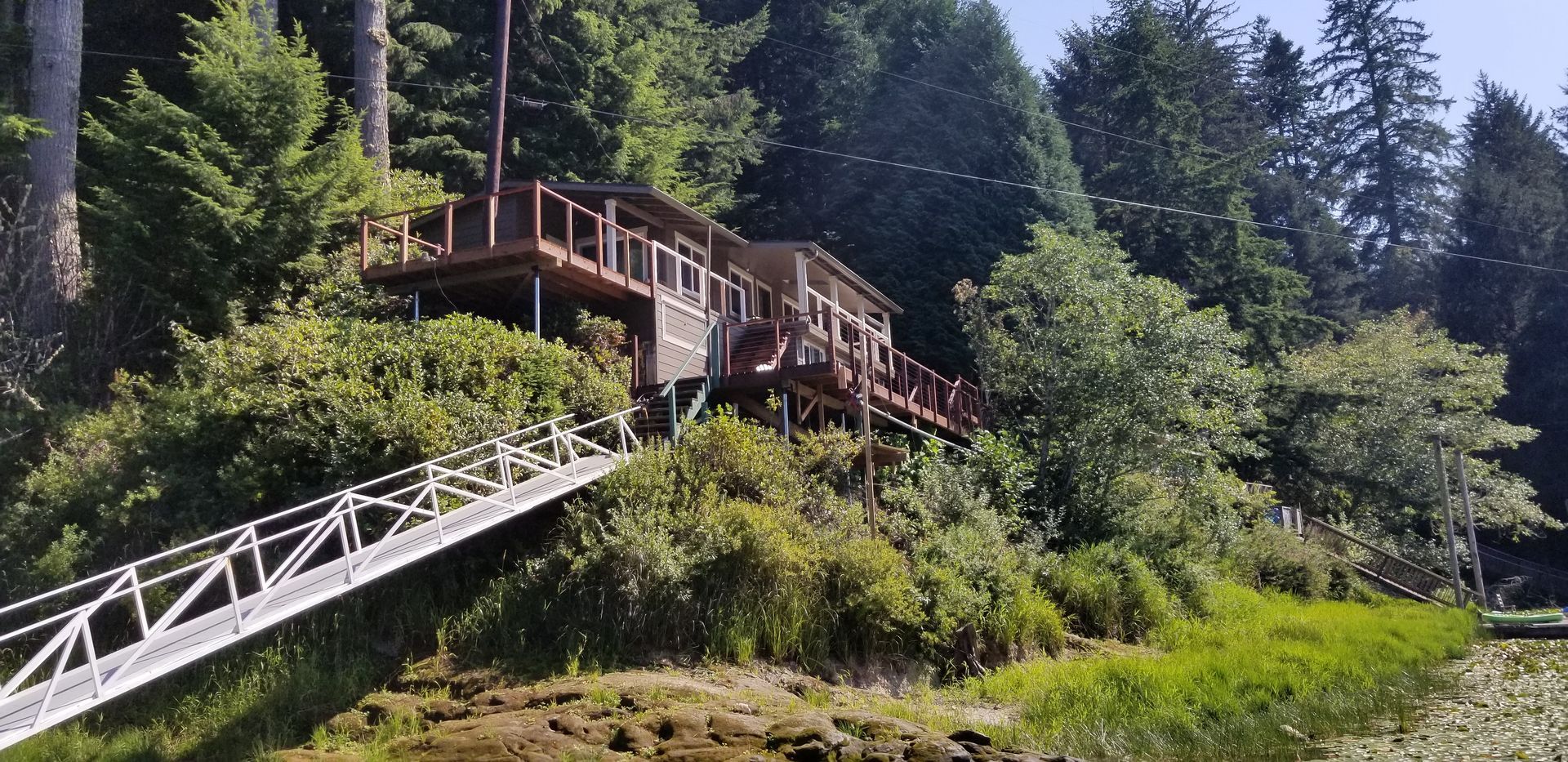 A wooden cabin with a ramp sits on a hillside, surrounded by lush green trees.