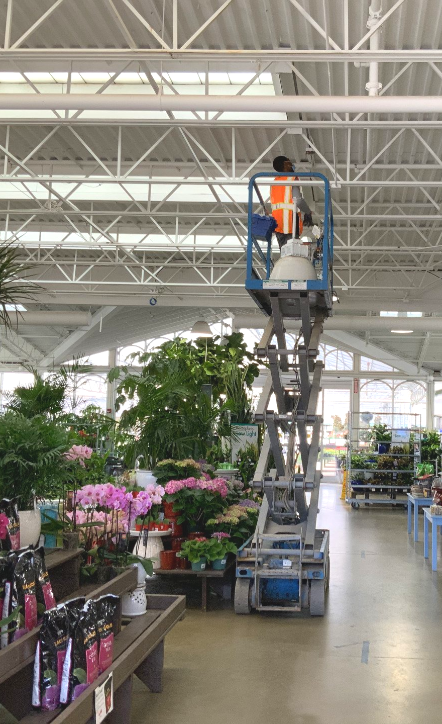 Worker in an orange vest on a raised scissor lift inside a bright greenhouse aisle