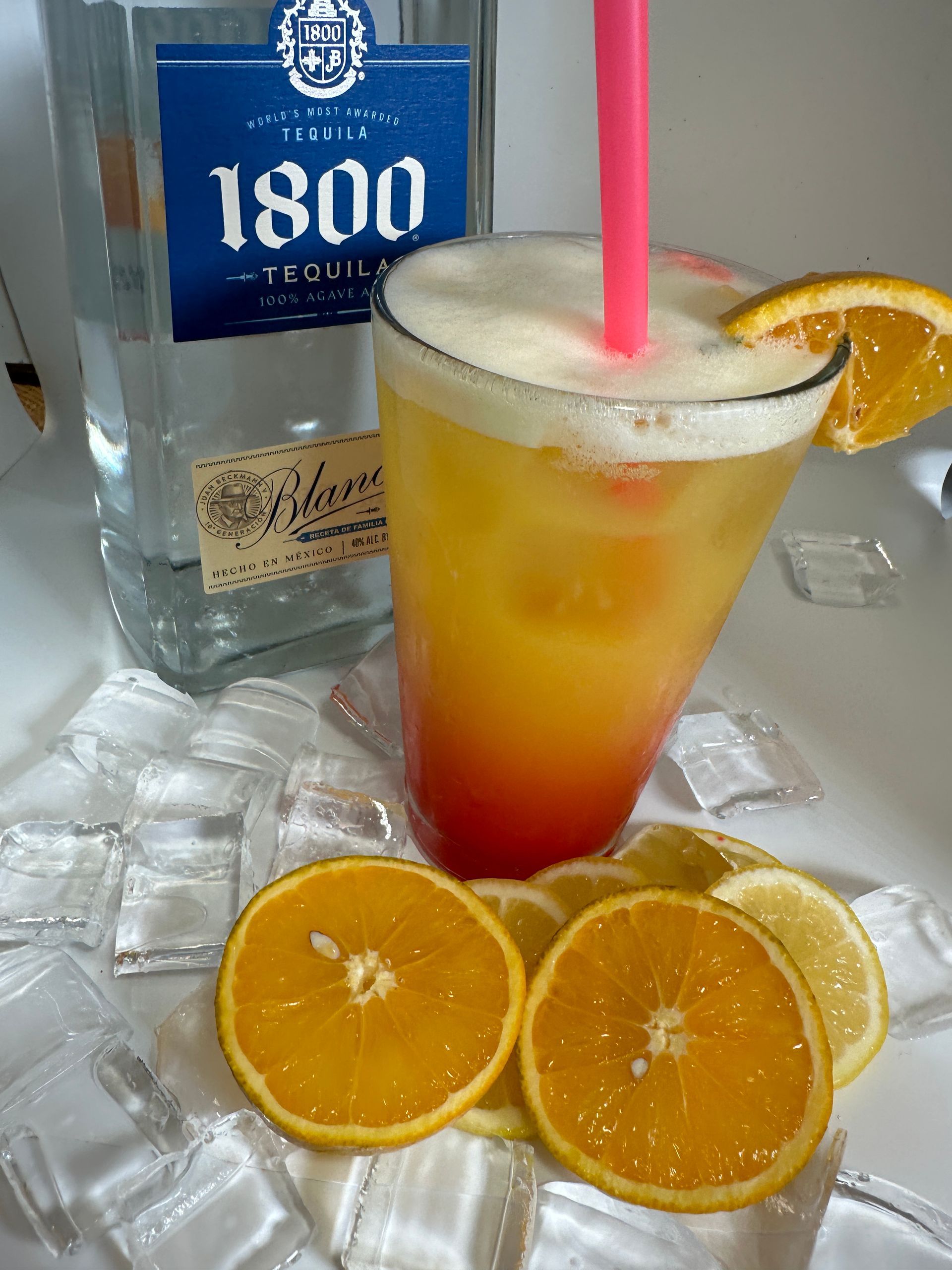 Cocktail with layered orange and yellow liquids, orange slice garnish, pink straw, bottle of tequila, orange and lemon slices.