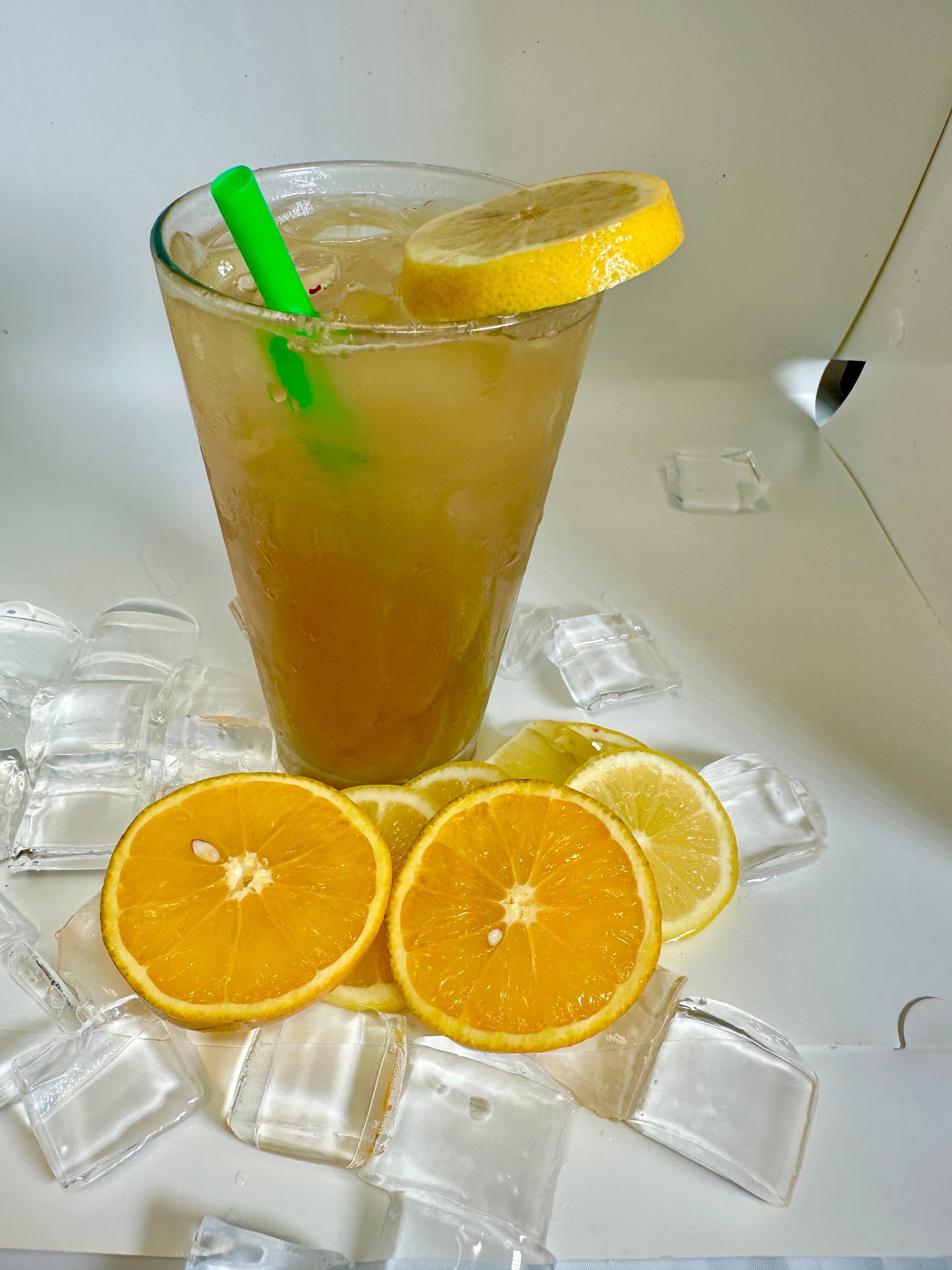 Tall glass of amber-colored drink with ice, green straw, lemon slice, surrounded by orange and lemon slices on ice cubes.