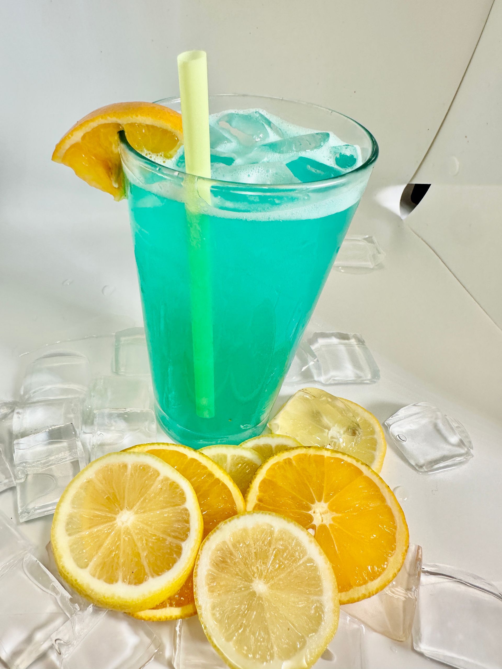 Blue cocktail in a glass with ice, orange slice, and straw, with lemon and orange slices nearby.