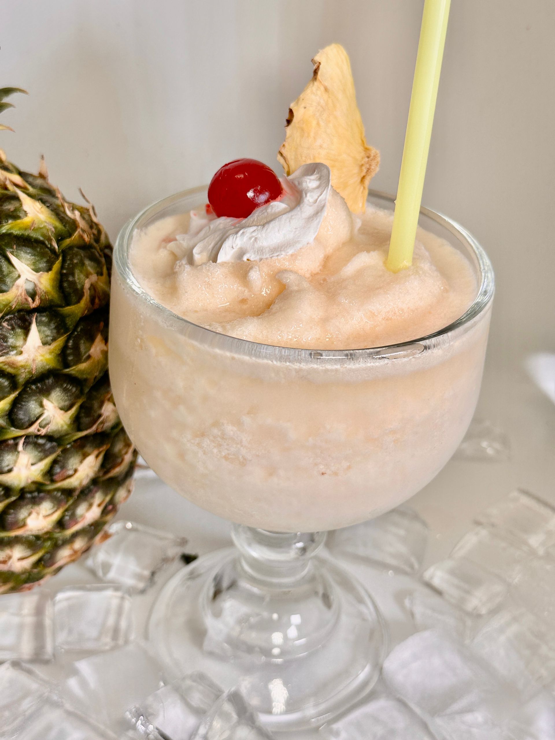 Pineapple cocktail in a stemmed glass, topped with whipped cream, cherry, pineapple slice, and straw, beside a pineapple.