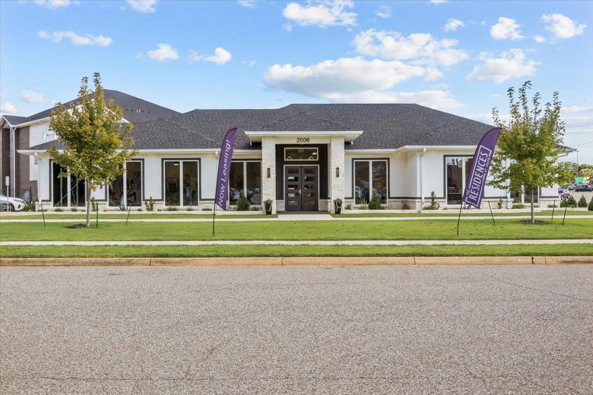 White building with black roof, glass windows, and purple flags on a lawn; a sunny day.