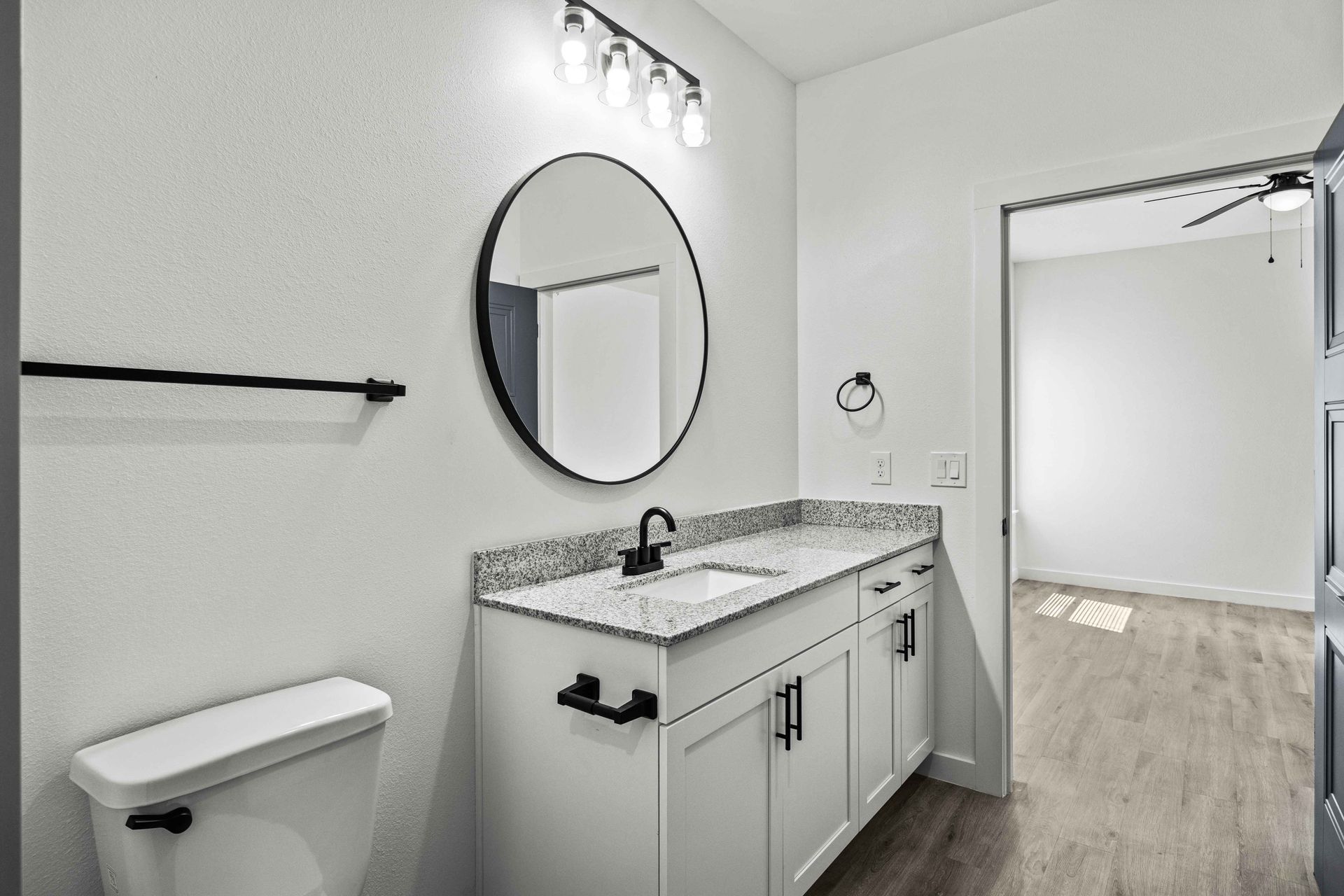White bathroom with vanity, toilet, round mirror, and doorway to another room.