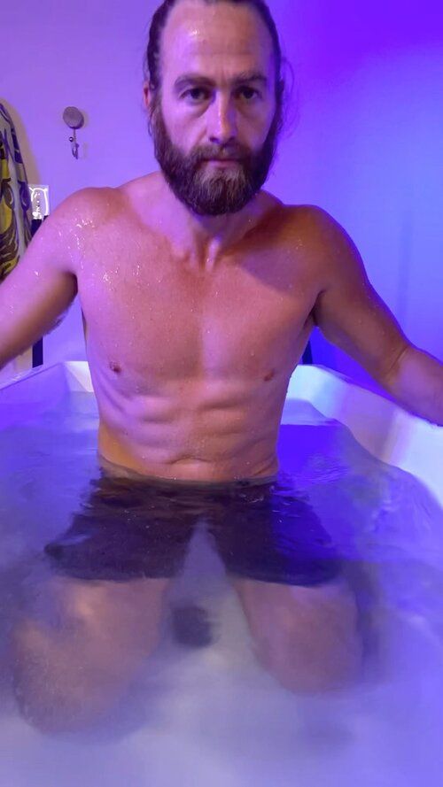 A shirtless man with a beard is sitting in a bathtub.