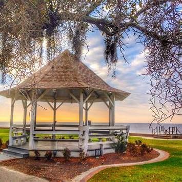 A white gazebo with a shingled roof sits on a green lawn overlooking a calm body of water at sunset.