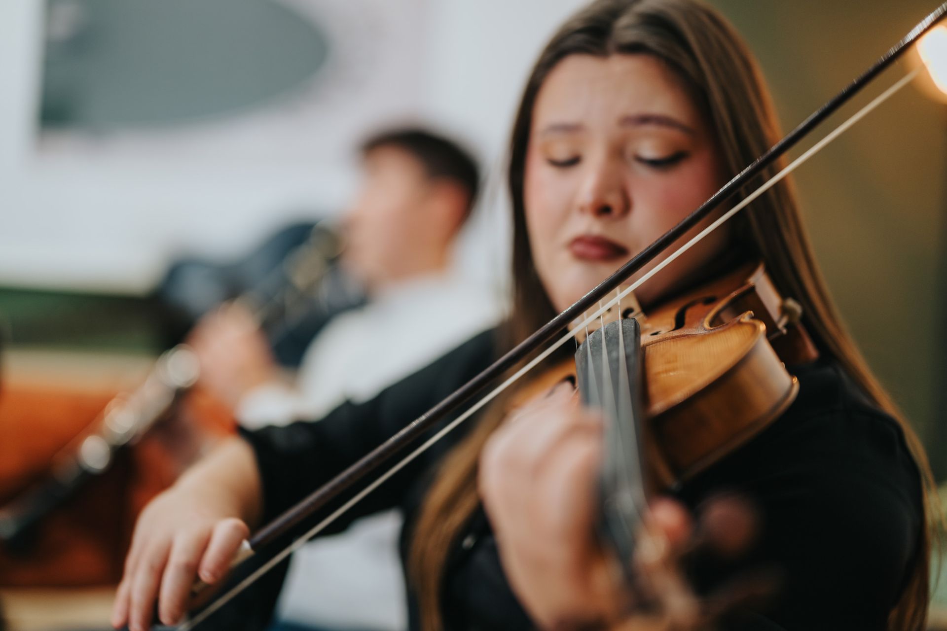 Woman playing a violin with a focused expression, another musician in the blurred background.