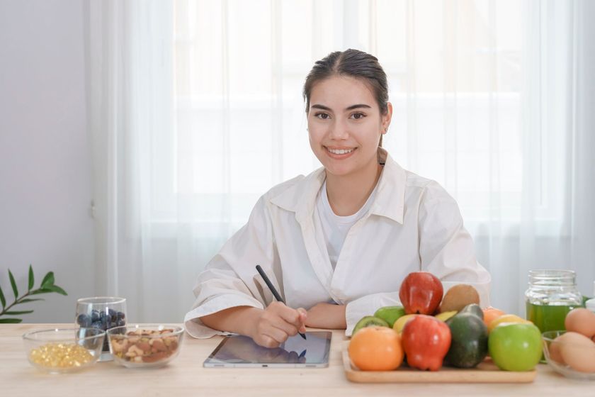 Woman smiling, drawing on tablet, surrounded by fruit, nuts, and supplements on a table. Woman smiling, drawing on tablet, surrounded by fruit, nuts, and supplements on a table.
