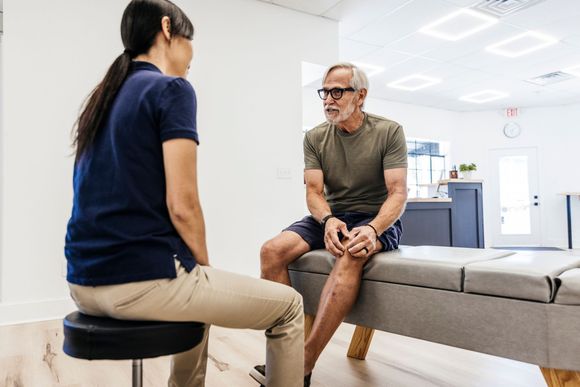 A therapist in a blue shirt talking with a patient seated on a therapy table, in a clinic room. A therapist in a blue shirt talking with a patient seated on a therapy table, in a clinic room.